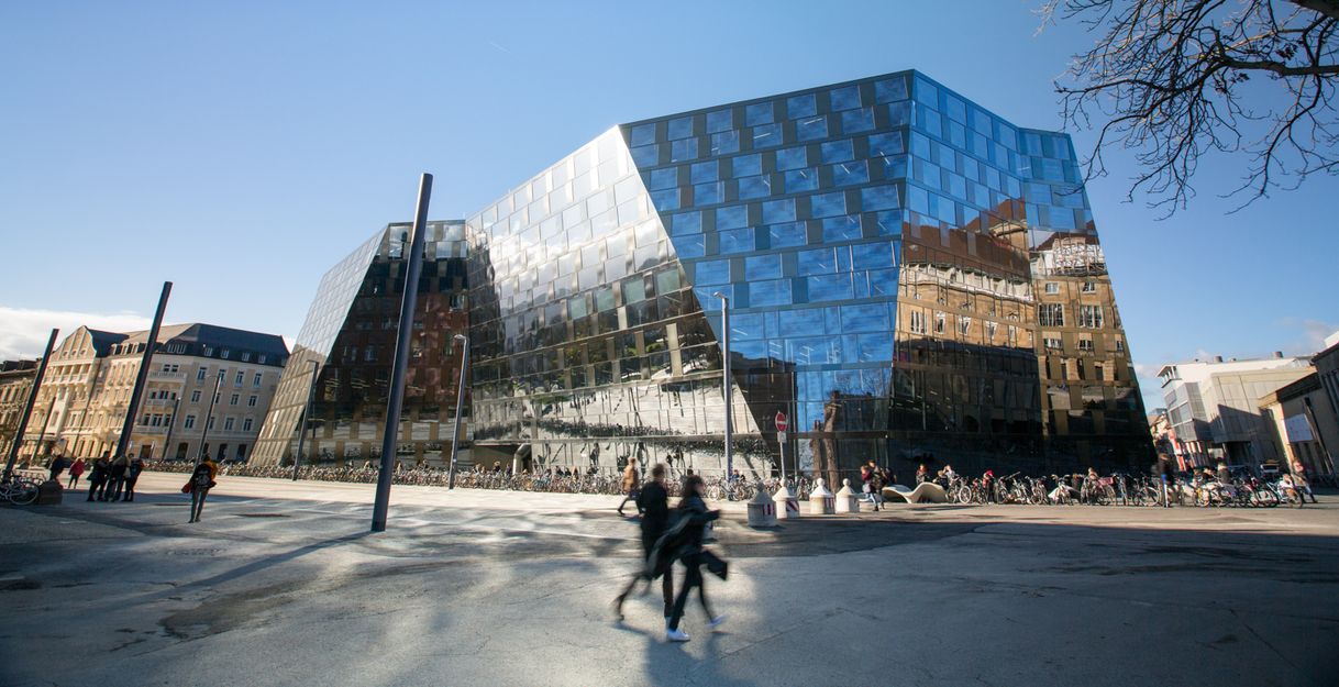 Das Bild zeigt das neue Gebäude vom Platz der Alten Synagoge fotografiert und mit einer gläsernen, verschieden geneigten Fassade. Der Himmel ist blau. Auf der Nordseite spiegelt sich das Stadttheater in der Fassade. Im Vordergrund laufen Menschen. 