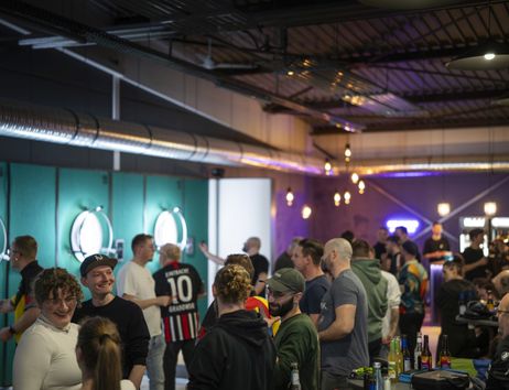 Dartzentrum Freiburg, Dartboards mit Beleuchtung an den Wänden, Bar und Lounge mit warmer Beleuchtung, Menschen, die Darts werfen, zuschauen oder miteinander sprechen. 