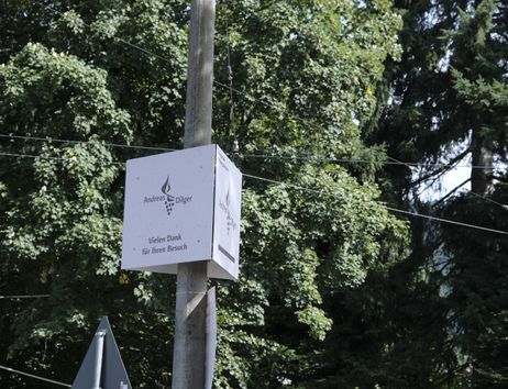 Ein Schild mit der Aufschrift 'Andreas Dilger' und 'Vielen Dank für Ihren Besuch' an einem Pfosten vor Bäumen.