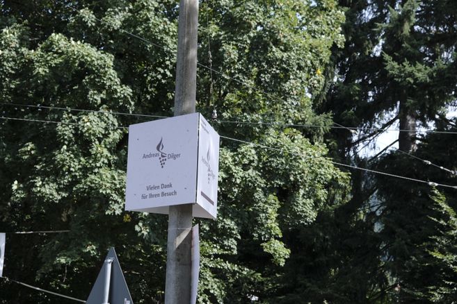 Ein Schild mit der Aufschrift 'Andreas Dilger' und 'Vielen Dank für Ihren Besuch' an einem Pfosten vor Bäumen.